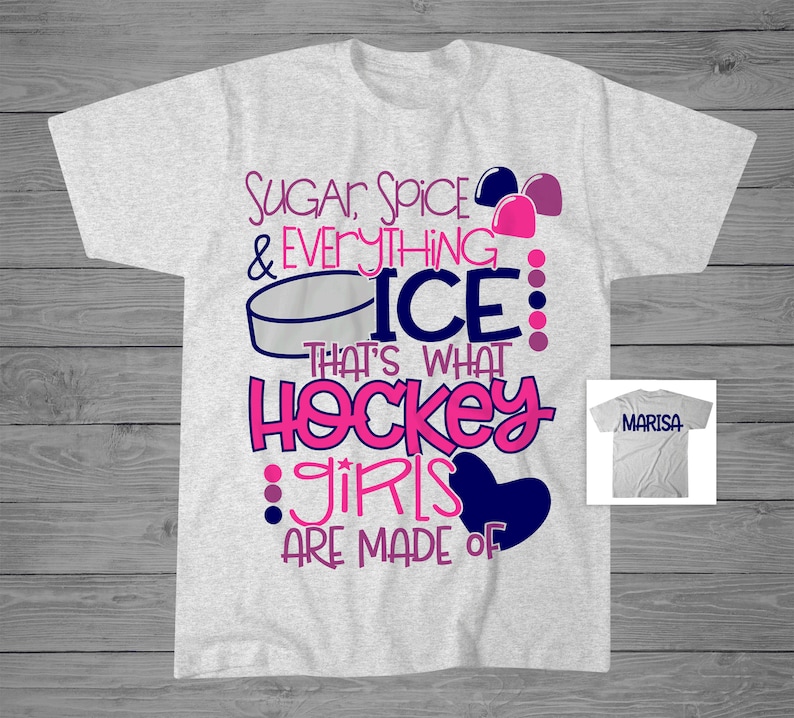 May include: A grey t-shirt with a pink and blue design that says "Sugar, Spice & Everything Nice: That's What Hockey Girls Are Made Of". The design also includes a hockey puck and a heart. The shirt is shown against a wooden background.