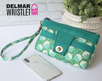 Pode incluir: Uma carteira de pulso turquesa com fecho de correr e uma aba com um fecho dourado. A carteira de pulso tem um padrão floral com detalhes em branco e dourado. A carteira de pulso está sobre uma superfície branca com um telefone branco e uma planta verde num vaso branco.