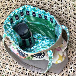 May include: A brown canvas tote bag with a zippered pocket and a green and blue geometric patterned lining. The bag is open and a black travel mug is inside.