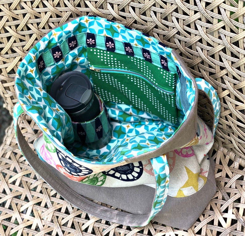 May include: A brown canvas tote bag with a zippered pocket and a green and blue geometric patterned lining. The bag is open and a black travel mug is inside.