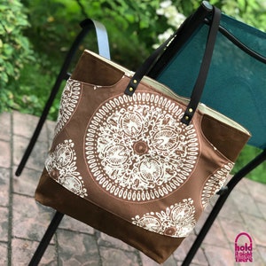 May include: A brown canvas tote bag with white mandala designs. The bag has black leather handles and a brown leather patch on the front. The bag is hanging on a chair.