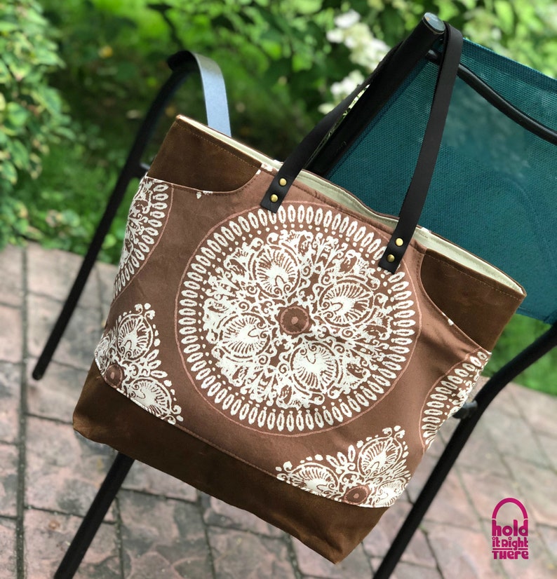 May include: A brown canvas tote bag with white mandala designs. The bag has black leather handles and a brown leather patch on the front. The bag is hanging on a chair.