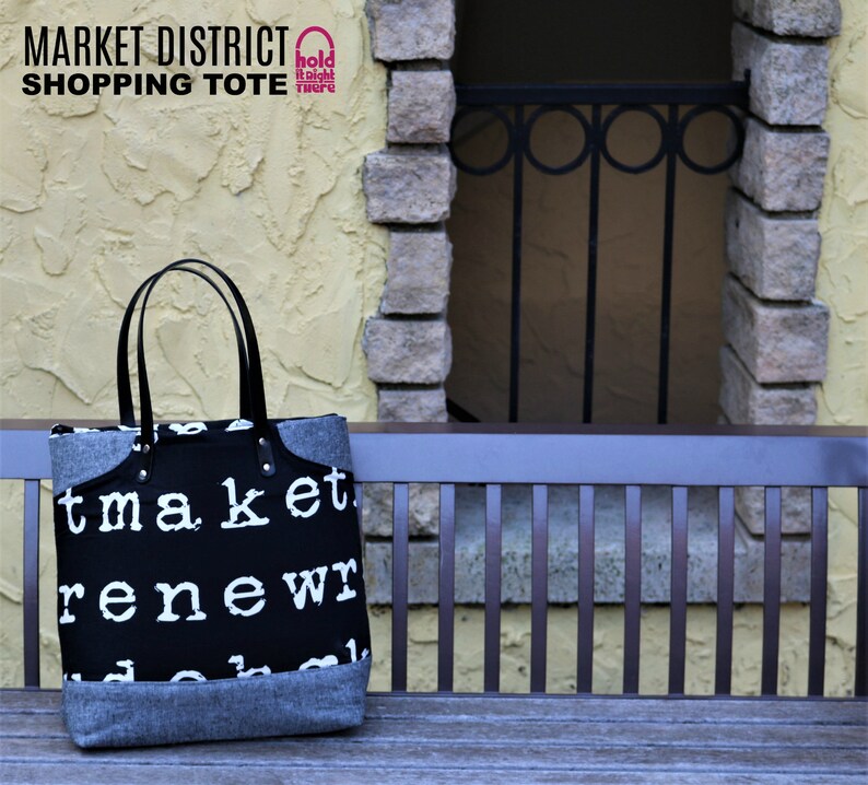 May include: Black and white shopping tote bag with the text "market renew" printed on it. The bag has black leather handles and a gray felt bottom. The bag is sitting on a wooden bench in front of a brick wall.