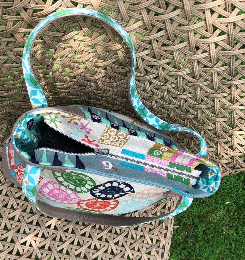May include: A colorful tote bag with a zipper closure and two handles. The bag is made of fabric with a patchwork design featuring geometric shapes, flowers, and other patterns. The bag is open and the inside is visible.