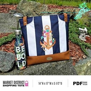 May include: A blue and white striped tote bag with a brown leather bottom. The bag features an embroidered anchor with floral accents. The bag is 14 inches high, 17 inches wide, and 5 1/2 inches deep. The text "MARKET DISTRICT SHOPPING TOTE" is printed on the bag.