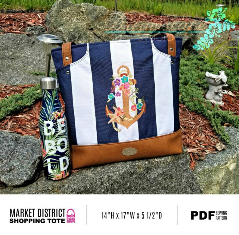 May include: A blue and white striped tote bag with a brown leather bottom. The bag features an embroidered anchor with floral accents. The bag is 14 inches high, 17 inches wide, and 5 1/2 inches deep. The text "MARKET DISTRICT SHOPPING TOTE" is printed on the bag.