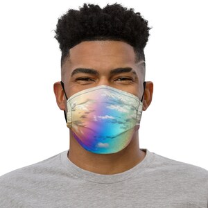 Puede incluir: Una mascarilla con un diseño de cielo y nubes. La mascarilla presenta un patrón iridiscente colorido con tonos de azul, amarillo y morado. La mascarilla tiene presillas negras y la lleva una persona.
