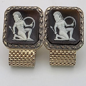 May include: A pair of vintage cufflinks featuring a square, dark brown cameo with a white classical figure. The gold-toned metal setting has a textured border and a mesh-like cuff. The cufflinks are displayed on a white background.