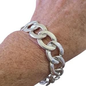 May include: A silver-colored chain link bracelet. The bracelet is made of large, rounded links. The bracelet is worn on a wrist, with the hand in the background. The bracelet is a bold, statement piece of jewelry.