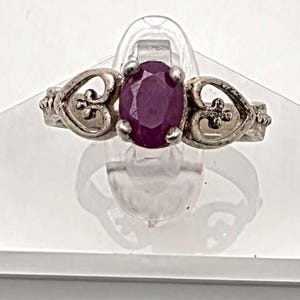 May include: A silver ring with a large oval ruby gemstone. The ring features decorative filigree hearts on each side of the ruby. The ring is displayed on a clear, angled stand against a white background.