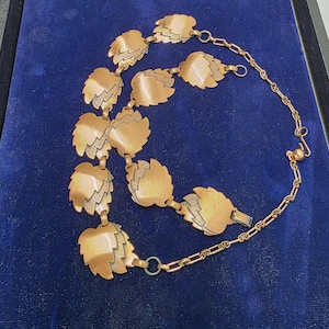 May include: A copper-toned necklace featuring leaf-shaped pendants with etched details. The necklace has a chain closure and is displayed on a blue velvet surface. The leaves are connected by small links, creating a unique and elegant design.