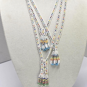 May include: Three beaded necklaces with white beads and colorful accents. Each necklace has a tassel pendant with a mix of colors, including blue, yellow, red, and green. The necklaces are displayed on a mannequin.