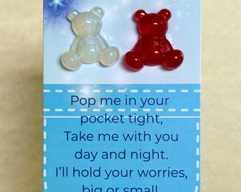 Op de afbeelding: Een kaart met twee kleine beer-vormige bedels, één doorzichtig en één rood, boven een blauw-witte kaart met de tekst: "Pop me in your pocket tight, Take me with you day and night. I'll hold your worries, big or small, And help you smile through it all."