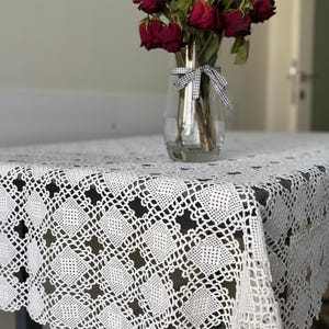 Puede incluir: Mantel de encaje blanco con un patrón cuadrado. Un jarrón con rosas rojas secas se encuentra sobre el mantel.