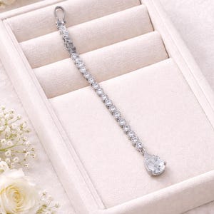 May include: A silver-coloured headpiece featuring a line of clear, round gemstones, ending in a teardrop-shaped pendant. It's presented on a white, velvet-lined jewellery box, with white flowers in the background.