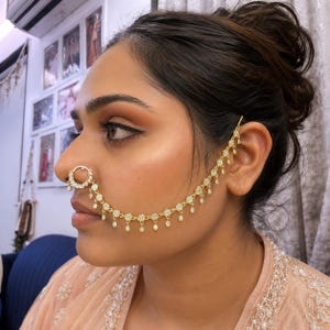 May include: A gold-toned nose ring connected to an ear cuff by a delicate chain. The nose ring features a circular design with small clear stones, while the chain consists of repeating floral-shaped links with dangling accents reaching the ear.