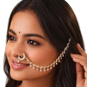 May include: A gold-toned nose ring connected to a delicate chain that extends towards the ear. The nose ring features a circular design encrusted with small clear stones, and the chain is adorned with multiple dangling teardrop-shaped crystals.