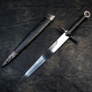 May include: A medieval-style sword with a silver blade and black handle, resting beside its black sheath. The handle features a decorative pommel and crossguard. The sheath has a textured design and silver accents.