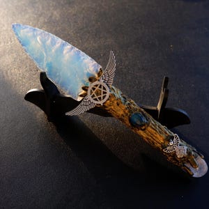 May include: A decorative dagger with a light blue, opalescent blade. The handle is textured with a brown and gold finish, adorned with silver-colored wings and a star emblem. A small owl emblem is at the base of the handle.