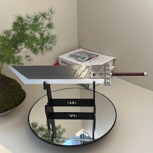 May include: A large, silver and gray Buster Sword replica with a brown handle, displayed on a black stand with a mirrored base. The sword has detailed metalwork and a wide blade, reminiscent of a video game weapon.