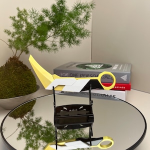 May include: A yellow, white, and black decorative knife with a circular handle, displayed on a black stand. The knife is resting on a reflective surface. The text "FOR ONE PERSON ONLY" is visible on a book in the background.
