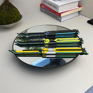 May include: Three butterfly knives with black, yellow, and teal accents are displayed on a round mirror. The knives have a sleek, modern design with angular blades and handles. The mirror reflects the knives and the surrounding objects.