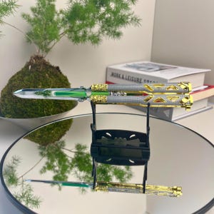 May include: A butterfly knife with a silver blade and handle, featuring gold and green accents. The knife is displayed on a black stand, reflecting on a mirrored surface. A small plant and books are in the background.