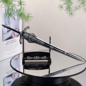 May include: A decorative sword with a black blade and ornate silver-colored hilt. The sword is displayed on a black stand, reflecting on a mirrored surface. The hilt features detailed carvings, and the blade has a central groove.