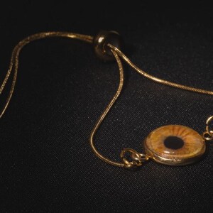 May include: A gold-toned bracelet featuring a circular pendant with an eye design. The eye has a brown iris and a black pupil. The bracelet has a delicate chain and a sliding bead for adjustment.