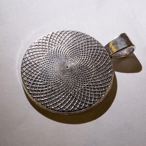 Op de afbeelding: Zilveren hanger met een cirkelvormig, geometrisch ontwerp. De hanger heeft een getextureerd oppervlak met een herhalend patroon van kleine vierkanten.