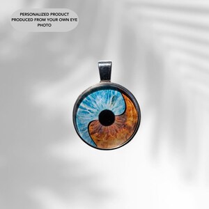 Op de afbeelding: Een zilveren hanger met een ronde glazen bedel met een blauw en bruin oogontwerp. Het oog is in twee&euml;n gedeeld, met een blauwe helft en een bruine helft.