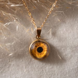 Pode incluir: Um colar com pendente dourado apresentando um berloque circular com um gráfico realista de um olho cor de âmbar. O olho tem uma pupila central escura rodeada por uma íris amarelo-dourada texturizada. O pendente está suspenso numa corrente dourada fina.