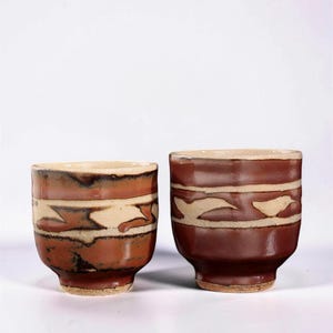 May include: Two ceramic cups with a reddish-brown glaze and a cream-colored band featuring abstract shapes. The cups have a slightly flared rim and a footed base. The design is repeated on both cups.
