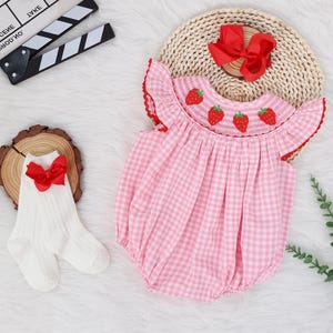 May include: Pink and white gingham baby onesie with embroidered strawberries. The onesie has a ruffled neckline and short sleeves.