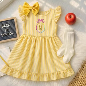 May include: Yellow striped dress with a bow, ruffle sleeves, and an embroidered initial. Includes white socks, a red apple, and a letter board that says "BACK TO SCHOOL". A pencil and notebook are also present.