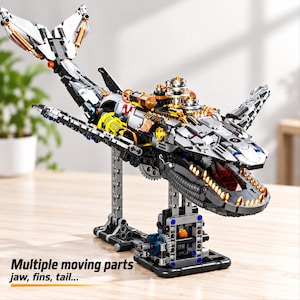 May include: A mechanical shark model constructed from interlocking building blocks, featuring a grey, black, and gold colour scheme with metallic accents. The model includes a detailed jaw with gold teeth, articulated fins, and a tail, mounted on a stand.