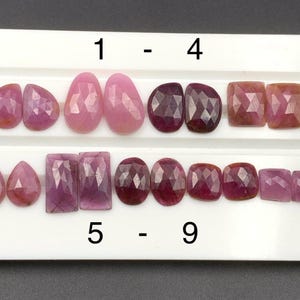 Könnte beinhalten: Auswahl an facettierten Edelsteinen in verschiedenen Formen und Farbtönen von Rosa und Rot, darunter ovale, tropfenförmige und quadratische Schliffe. Die Edelsteine sind auf einer weißen Präsentationsfläche angeordnet, mit den Zahlen 1-9.