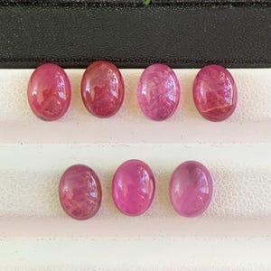 May include: Eight oval-shaped gemstones in varying shades of pink and red. The stones are polished and have a translucent appearance, with internal patterns visible. The gemstones are arranged in two rows against a neutral background, showcasing their color variations.