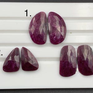 May include: Three pairs of faceted, oval, and rectangular-shaped gemstones in varying sizes. The gemstones are a deep, rich magenta color with a sparkling, textured surface. The gemstones are displayed on a white surface.