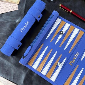 Puede incluir: Un juego de backgammon azul enrollable con el nombre Phoebe impreso en blanco. El tablero incluye puntos triangulares blancos y color canela, un compartimento de almacenamiento con cremallera y dos dados blancos. Incluye un estuche cilíndrico.