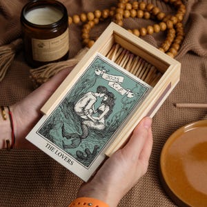 May include: A rectangular wooden matchbox featuring a sliding cover with a green and black illustration of a mermaid and a person embracing. The text reads THE LOVERS, 2025, and K+L. The box is filled with wooden matches with light-coloured tips.