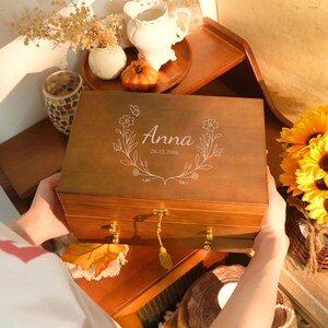 Könnte beinhalten: Eine rechteckige Schmuckschatulle aus Holz mit Klappdeckel, verziert mit einem weißen gravierten Blumenkranz, dem Namen Anna und dem Datum 26.12.2006. Die Schatulle hat ein goldenes Schlüsselloch mit Quastenschlüssel und kleine goldene Knöpfe.