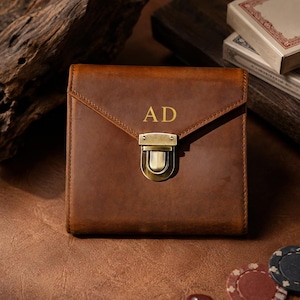 Puede incluir: Una cartera cuadrada de cuero marrón con un cierre de presión de metal dorado y las letras AD grabadas en la solapa frontal. La cartera tiene un acabado mate liso y un diseño estructurado tipo sobre con costuras visibles en los bordes.