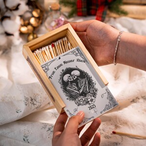 Puede incluir: Una caja de cerillas de madera con una tapa corredera decorativa. La tapa muestra el texto "True Love Never Dies" y una ilustración de dos esqueletos abrazándose. La caja contiene cerillas.