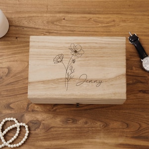 May include: A light-colored wooden box with a floral design and the name "Jenny" engraved on the lid. The box is rectangular with a small pull tab on the front. A watch and a strand of pearls are visible.