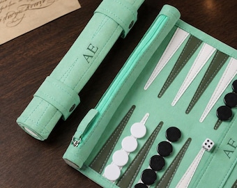 May include: A mint green roll-up backgammon set featuring the letters AE embossed on the exterior case and the interior board. The set includes white and black circular checkers, dice, and a matching mint green zippered storage pouch for pieces.
