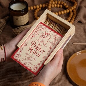 May include: A rectangular wooden matchbox featuring a red and cream label with an illustration of a cupid, floral scrollwork, and the text "The Perfect Match," "Made for Each Other," "Joe & Luna," and "2026." The box contains wooden matches.