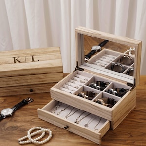 Può includere: Un portagioie in legno chiaro con coperchio a specchio, aperto per rivelare scomparti per anelli e orologi. Un cassetto sottostante contiene collane. È visibile anche una seconda scatola con le iniziali "K|L".