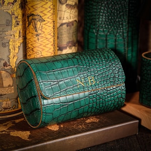 May include: A green, crocodile-textured watch case with gold-colored initials "NB" on the front. The case is cylindrical with a flap closure and brown trim. The background includes antique books and a world map.