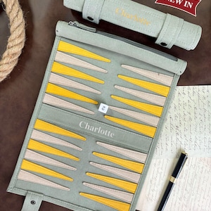 May include: A sage green roll-up backgammon board featuring yellow and beige triangular points, the name Charlotte printed in the center, a small white die with the number 2, and a matching sage green storage case with the text NEW IN.